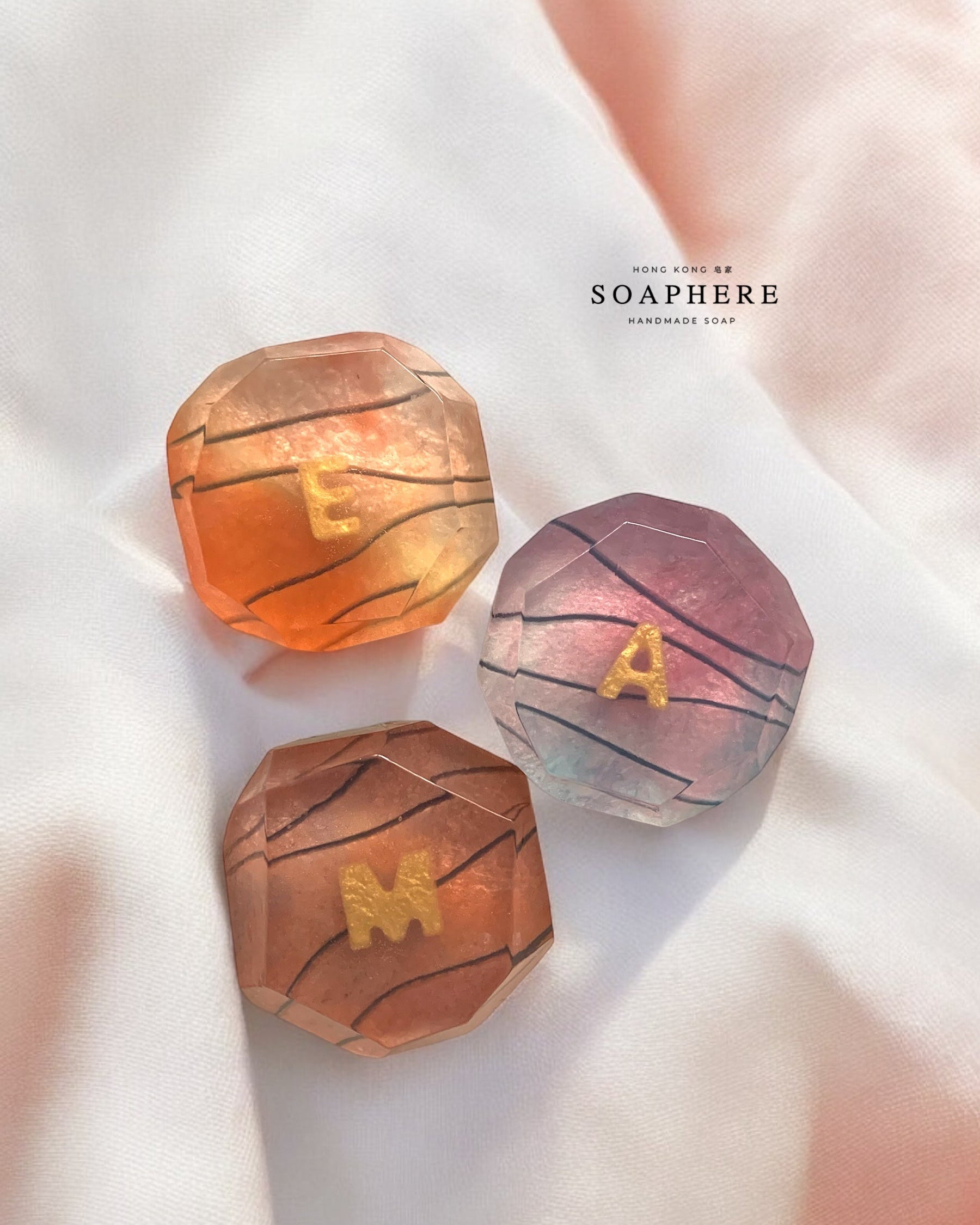Three colorful geometric stone soaps with letters on a soft pink background.